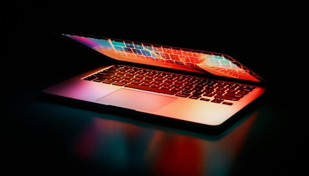 Laptop partially opened, emitting vibrant multicolored lights in dark setting.